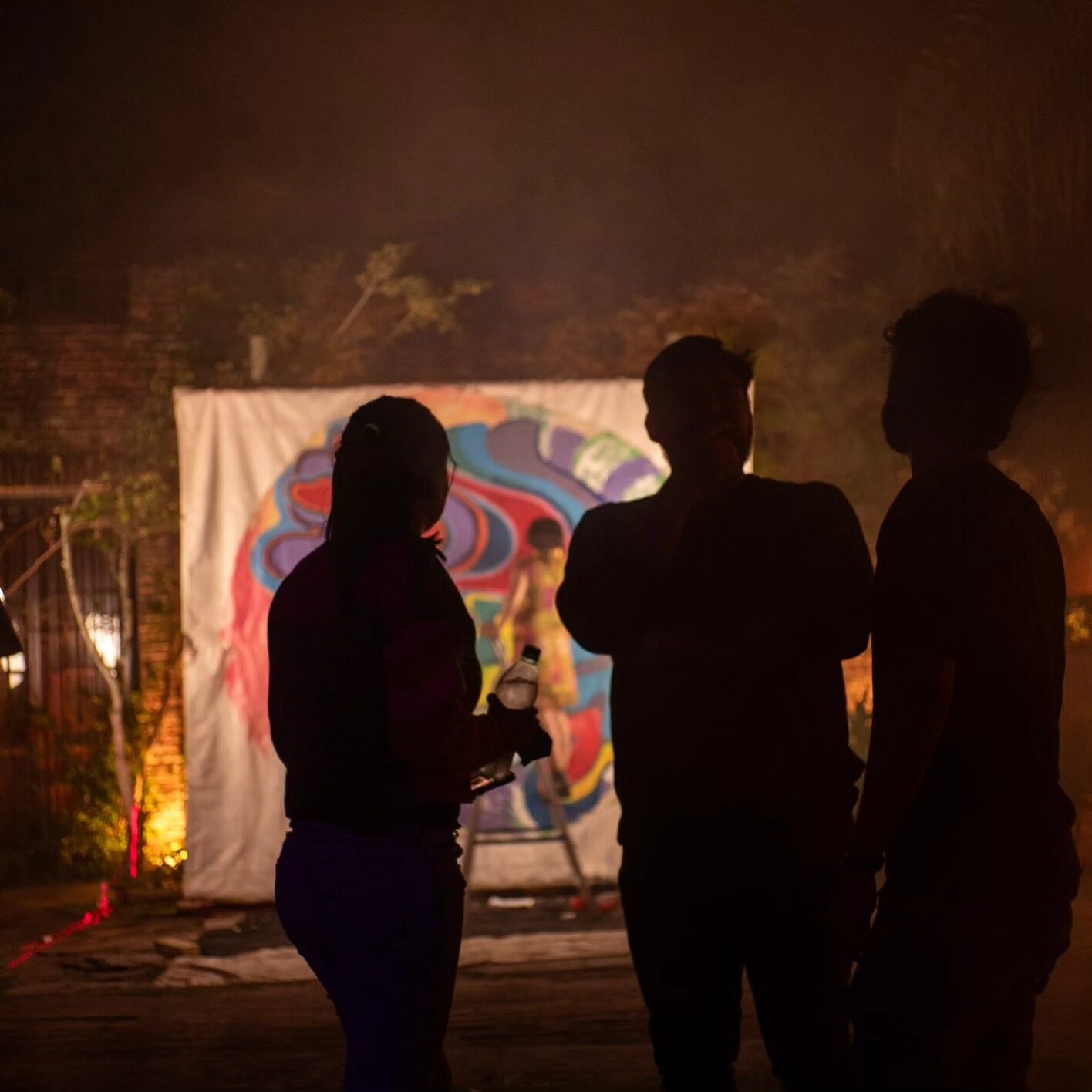 Tres siluetas frente a un lienzo pintado e iluminado, de noche.
